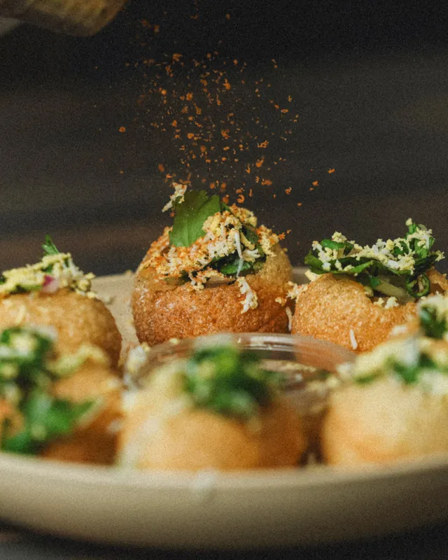 Fuchka shells topped with herbs and grated egg, with chili powder falling over the plate