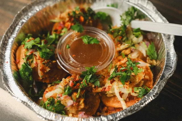 A takeout tray of fuchka loaded with onion, cilantro and chili flakes around a cup of tamarind water